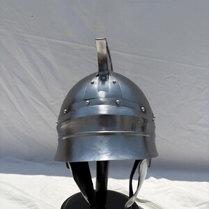 May include: A polished silver-colored metal helmet featuring a rounded dome, a central vertical crest, and a wide brim. The helmet is constructed with visible rivets along the crown and includes dark leather chin straps hanging from the interior.