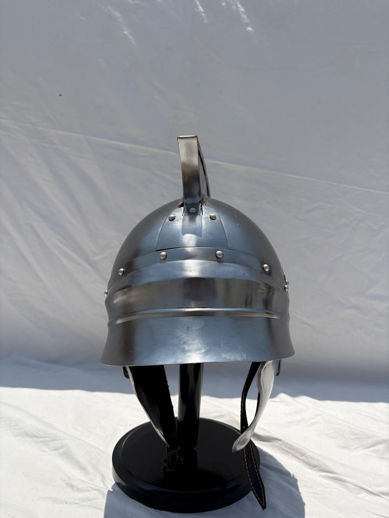 May include: A polished silver-colored metal helmet featuring a rounded dome, a central vertical crest, and a wide brim. The helmet is constructed with visible rivets along the crown and includes dark leather chin straps hanging from the interior.