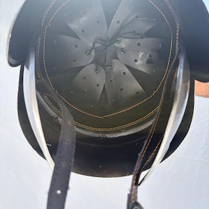 May include: A dark-colored protective helmet viewed from the interior, featuring a grey multi-panel suspension liner with ventilation holes, secured by a black leather chin strap with orange stitching and a metal buckle attached to the rim.