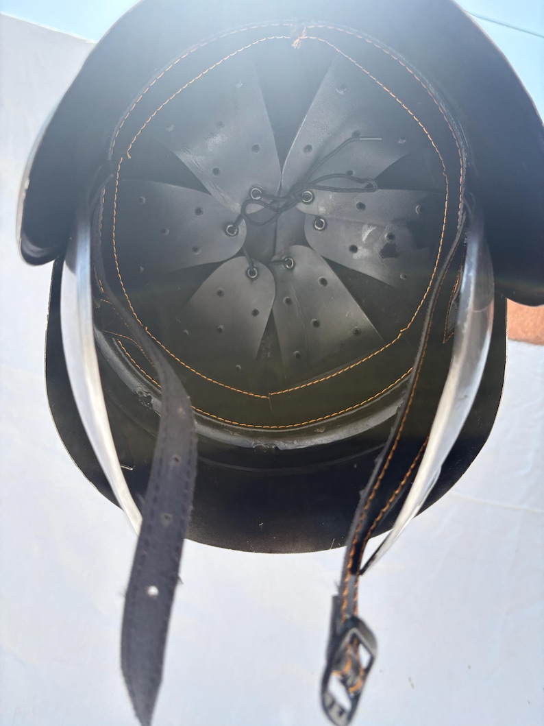 May include: A dark-colored protective helmet viewed from the interior, featuring a grey multi-panel suspension liner with ventilation holes, secured by a black leather chin strap with orange stitching and a metal buckle attached to the rim.