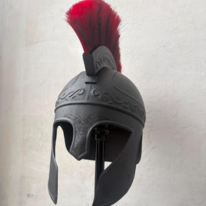 May include: A matte black helmet featuring a prominent red crest made of coarse hair. The helmet displays embossed scrollwork patterns along the crown and a stylized animal head design centered above the face opening, providing full coverage.