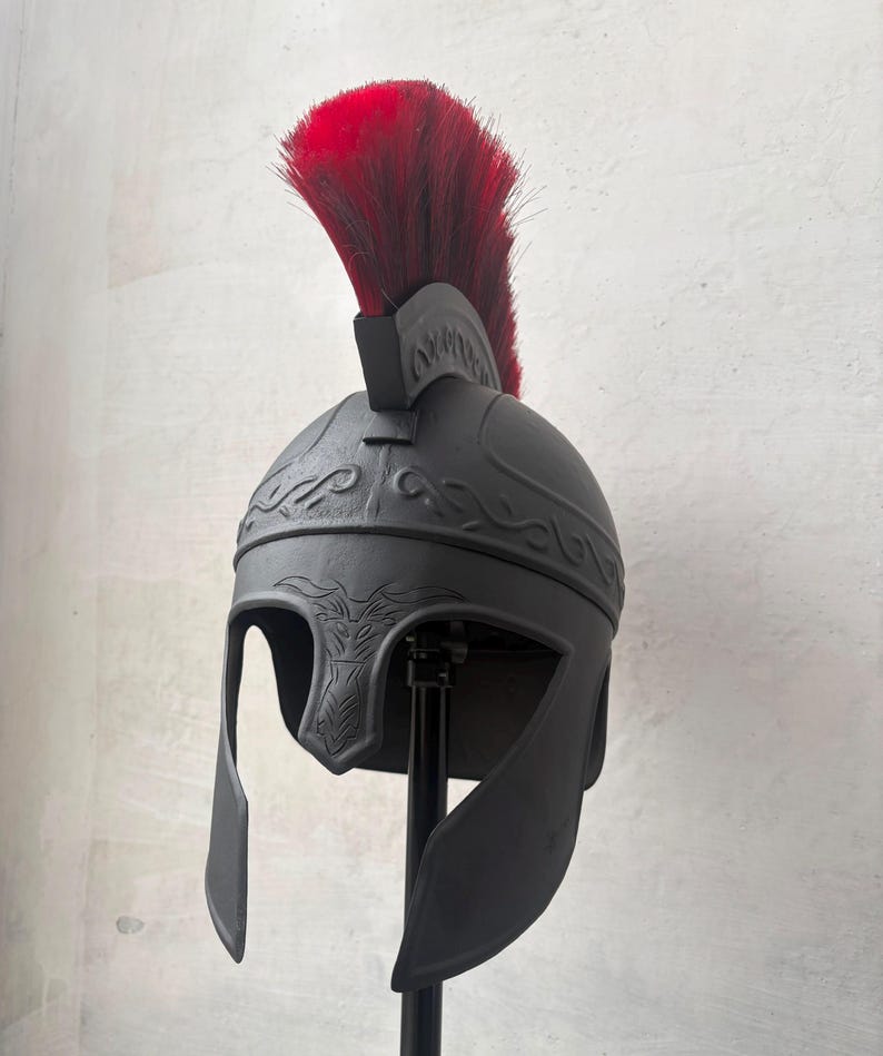 May include: A matte black helmet featuring a prominent red crest made of coarse hair. The helmet displays embossed scrollwork patterns along the crown and a stylized animal head design centered above the face opening, providing full coverage.