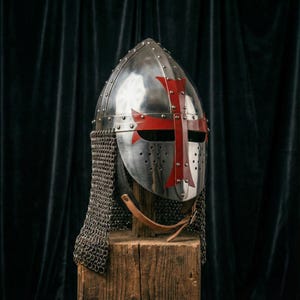 Può includere: Un elmo in metallo lucido con un design a croce rossa, con una protezione per il collo in cotta di maglia. L'elmo è montato su un blocco di legno contro uno sfondo di tenda scura. C'è un cinturino in pelle.