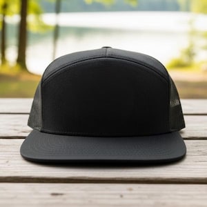 May include: A black flat-peak cap with a mesh back. The cap features a structured front panel and a flat peak. The mesh back offers ventilation. The cap is on a wooden surface, with a blurred background of trees and water.