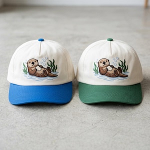 Op de afbeelding: Twee baseballpetten met een crèmekleurige kroon en een gekleurde klep. Eén heeft een blauwe klep en de andere een groene klep. Elke pet heeft een geborduurd otterontwerp, waarbij de otter in het water drijft.