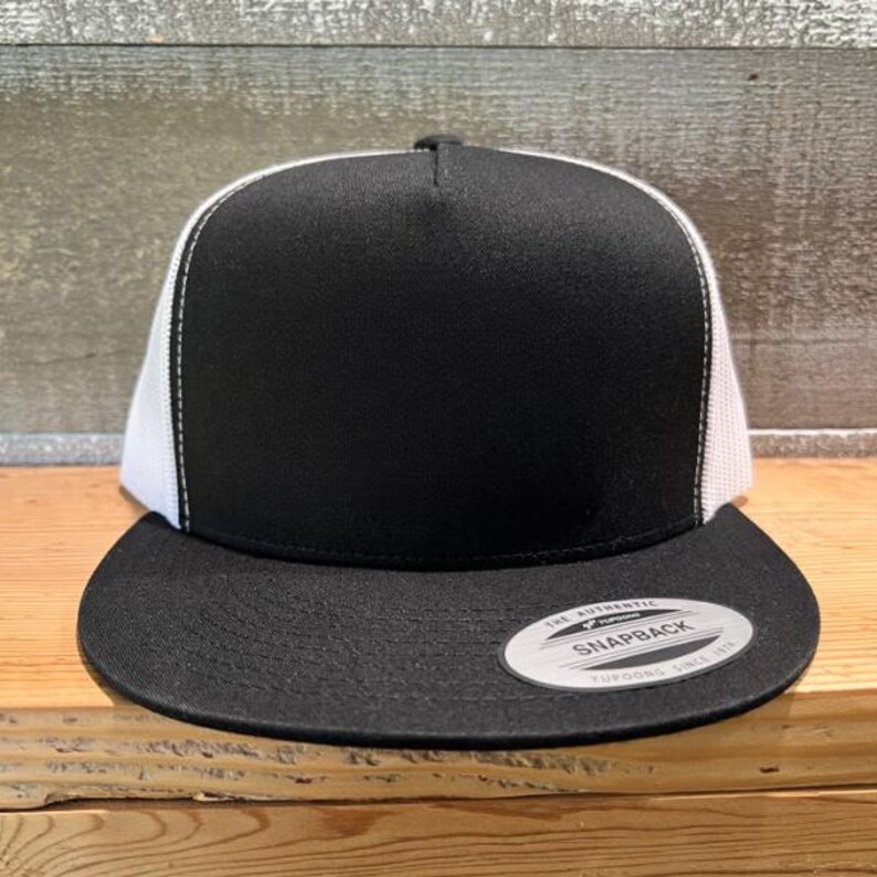 May include: A black and white trucker cap with a flat peak. The front panel is black, and the mesh back is white. A silver and black circular tag is on the front of the peak. The cap sits on a wooden surface.