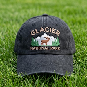 May include: A black baseball cap featuring an embroidered design of white mountains, green pine trees, and a brown elk. The text GLACIER NATIONAL PARK is stitched in tan thread above and below the central mountain and elk graphic.