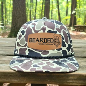 Puede incluir: Gorra de camionero con estampado de camuflaje y parche de cuero marrón. El parche presenta la palabra "BEARDED" encima de "FOR HER PLEASURE" y una ilustración de un vikingo. El sombrero tiene un detalle de cuerda negra a lo largo del borde.