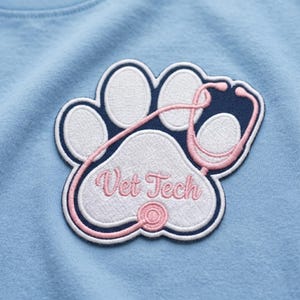 May include: A light blue t-shirt featuring an embroidered patch shaped like a paw print. The patch has a dark blue background with white paw pads and a pink stethoscope outline. The text Vet Tech is embroidered in pink cursive script.