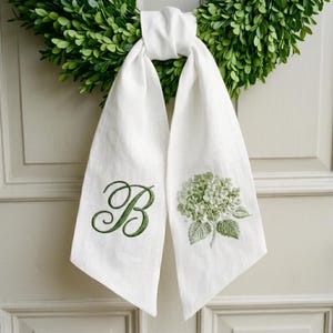 May include: A white fabric sash featuring an embroidered green letter B on one side and a detailed green hydrangea flower illustration on the other. The sash is tied in a knot at the top and hangs down in two long, pointed ends against a green wreath.