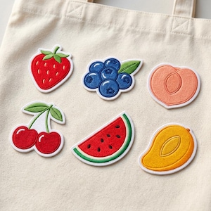 May include: Embroidered fruit patches including a red strawberry, blueberries, a peach, cherries, a watermelon slice, and a mango. Each patch has a white border and is displayed on a beige tote bag. These patches are ideal for adding a touch of summer to clothing or accessories.