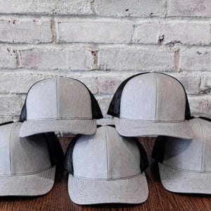 May include: A collection of six gray and black trucker hats. The hats have a gray front panel and brim, with a black mesh back. They are arranged on a wooden surface in front of a brick wall.