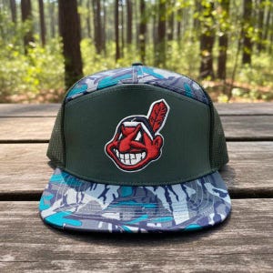May include: A camouflage-patterned trucker hat with a green front panel featuring a red and white cartoon logo. The hat has a mesh back and a curved brim. The camouflage pattern includes shades of blue, gray, and white.
