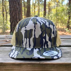 May include: A camouflage print trucker cap with a curved peak. The hat has a mesh back and a front panel with a green, white, and navy blue camouflage design. A small, circular tag is on the front.