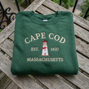 Puede incluir: Una sudadera verde oscuro con el texto bordado "CAPE COD" sobre un dibujo de un faro. Debajo del faro están las palabras "EST. 1637" y "MASSACHUSETTS". La sudadera está doblada sobre una superficie de madera.