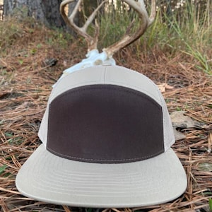 May include: A tan and brown trucker hat with a mesh back and a flat peak. The front panel is brown, and the peak and top are tan. The hat is on a bed of pine needles.