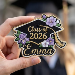 Op de afbeelding: Een zwarte afstudeerpet-patch met gouden rand, met de tekst "Class of 2026 Emma". De patch is versierd met geborduurde paarse en witte bloemen en een gouden kwast, perfect om afstuderen te vieren.