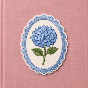 May include: An oval-shaped embroidered patch featuring a blue hydrangea flower with green leaves on a white background. The patch is framed by a scalloped light blue border with detailed stitching, creating a decorative floral design element.