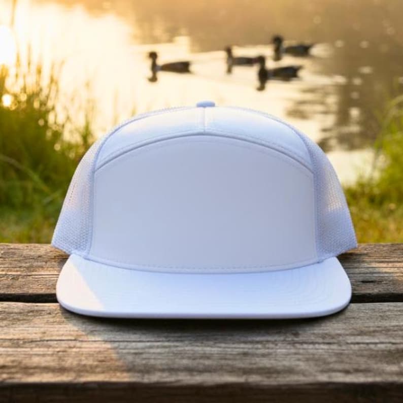 May include: A white trucker cap with a flat peak, featuring a solid front panel and mesh side and back panels. The hat is set against a blurred background of a lake and greenery, resting on a wooden surface.
