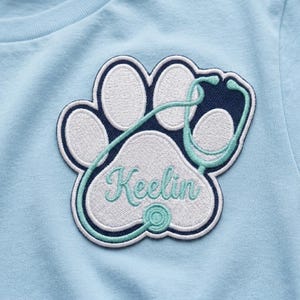 May include: A light blue t-shirt featuring a paw print design with a stethoscope and the name "Keelin" embroidered in a script font. The paw print is white with a navy blue outline, and the stethoscope is teal.