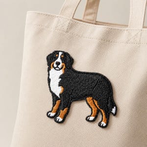 May include: A beige canvas tote bag featuring a detailed embroidered patch of a Bernese Mountain Dog. The dog is depicted in its characteristic black, white, and tan coat, standing in profile with its head turned toward the viewer.