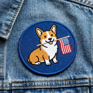 May include: Embroidered patch featuring a corgi dog holding a small American flag in its mouth. The patch is round with a blue background and a blue border. The corgi is brown, white, and tan.