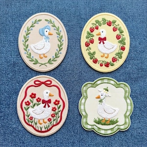 Op de afbeelding: Vier geborduurde patches met ganzenontwerpen. Eén toont een gans met een blauwe muts, een andere met een rode strik en aardbeien, een derde met rode bloemen en de laatste met een bloemenkrans.