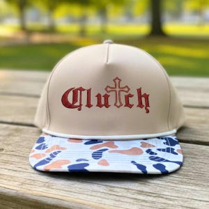 May include: A beige baseball cap with the word "Clutch" in red gothic lettering and a cross design. The brim features a camouflage pattern in blue, orange, and white.