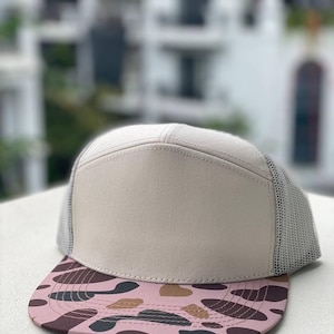 May include: A beige trucker cap with a mesh back and a flat peak. The peak has a pink and brown camouflage design. The front panel of the cap is a solid beige, and the mesh is light grey.