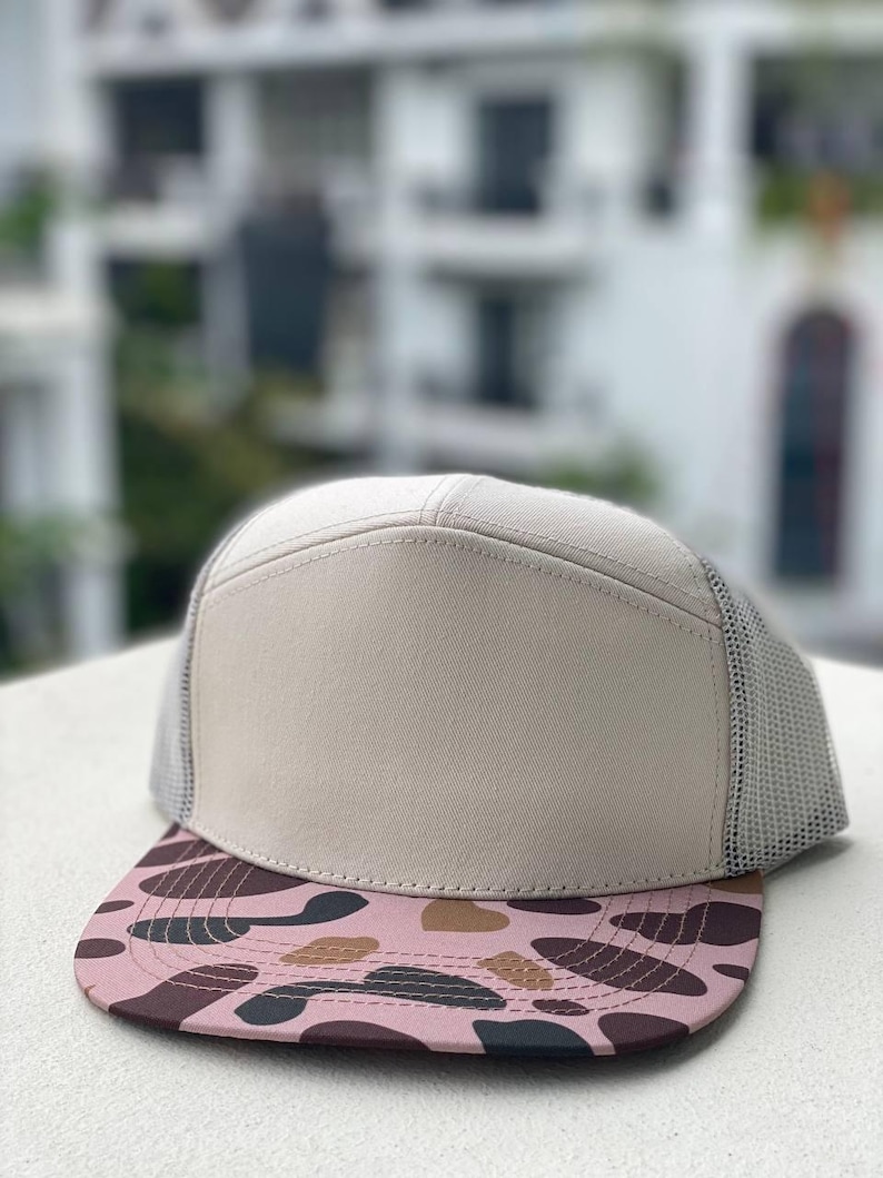May include: A beige trucker cap with a mesh back and a flat peak. The peak has a pink and brown camouflage design. The front panel of the cap is a solid beige, and the mesh is light grey.