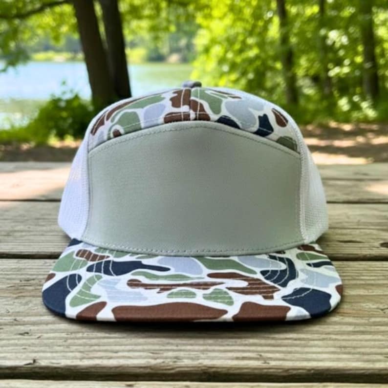 May include: A flat-peak cap featuring a camouflage design on the peak and crown. The front panel is a solid light grey, and the sides are white mesh. The cap is resting on a wooden surface.