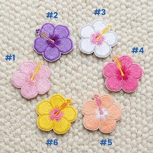 May include: Six embroidered hibiscus flower patches in various colors: pink, purple, white, yellow, and peach. Each patch has a detailed design with a yellow stamen and a white border. The patches are numbered from 1 to 6.