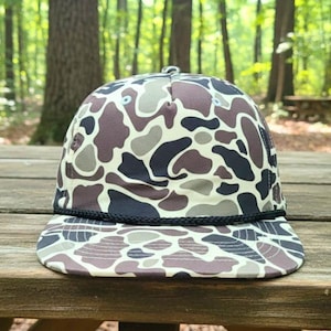 May include: A flat-brimmed baseball cap with a camouflage design. The cap has a black rope detail on the brim and a pattern of brown, grey, and off-white shapes. The background is a blurred view of a forest.