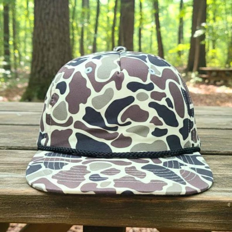 May include: A flat-brimmed baseball cap with a camouflage design. The cap has a black rope detail on the brim and a pattern of brown, grey, and off-white shapes. The background is a blurred view of a forest.