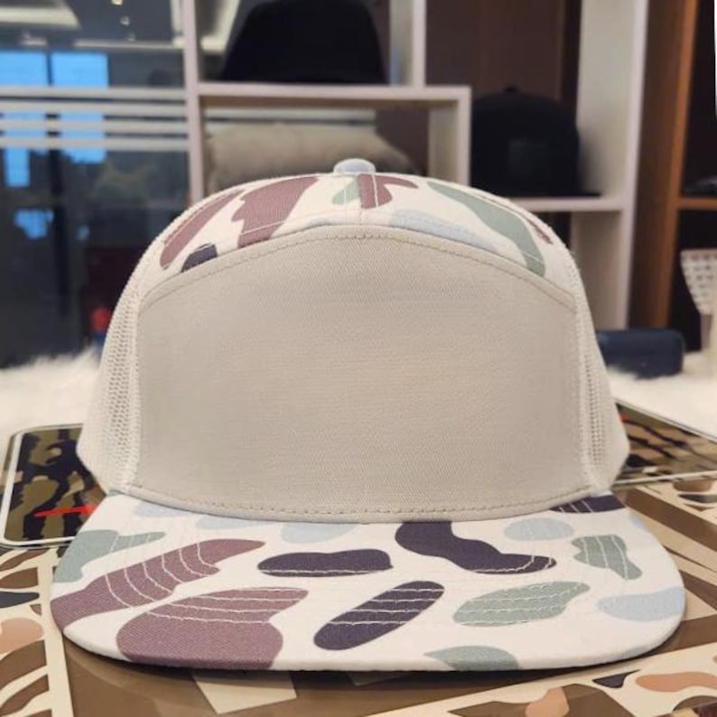 May include: A beige and white camouflage print baseball cap with a mesh back. The cap has a flat peak with a pattern of purple, green, and grey shapes. The front panel is a solid beige colour.