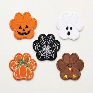 May include: Five embroidered paw-shaped patches featuring seasonal designs: an orange jack-o'-lantern, a white ghost, a black spiderweb, an orange pumpkin with a green stem, and a brown turkey with a yellow beak and red wattle on a white background.