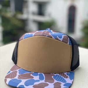 May include: A flat-brimmed cap featuring a brown front panel and a blue, brown, and white camouflage pattern on the brim and top. The sides and back are black mesh. The hat is on a white surface.