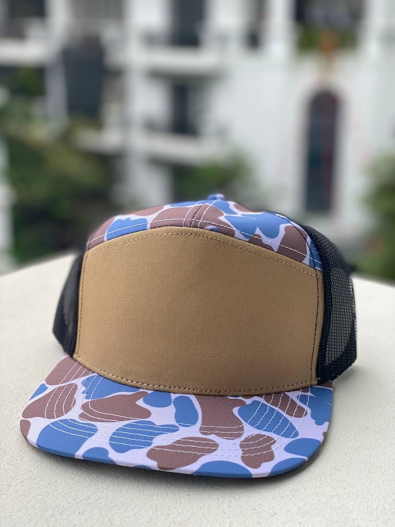 May include: A flat-brimmed cap featuring a brown front panel and a blue, brown, and white camouflage pattern on the brim and top. The sides and back are black mesh. The hat is on a white surface.