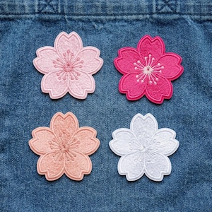 May include: Four embroidered flower patches in shades of pink and white. The patches are in the shape of cherry blossoms, with detailed stitching. The patches are arranged on a blue denim background.