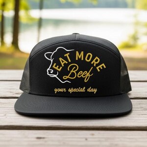 May include: Black trucker hat with a flat peak and mesh back. The front displays the phrase "EAT MORE Beef" in gold and white, with a cow graphic. The text "your special day" is below the main text.