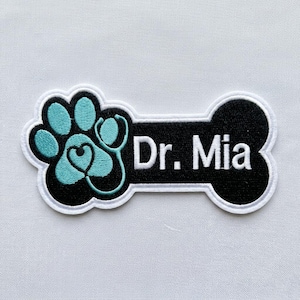 May include: Embroidered patch featuring a teal paw print with a stethoscope forming a heart shape, and the text "Dr. Mia" on a black bone-shaped background, outlined in white.