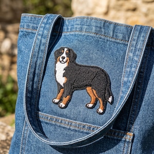 May include: A blue denim tote bag featuring an embroidered patch of a Bernese Mountain Dog. The dog is black, white, and tan, with a detailed design. The bag has denim straps and a pocket.