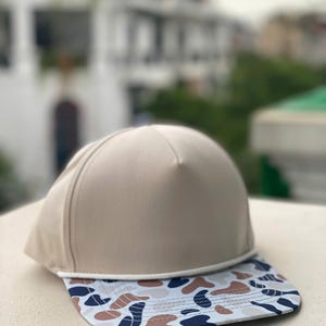 May include: A beige baseball cap with a flat peak. The peak has a pattern of blue, brown, and white shapes. The cap is made of a smooth material and has a classic, sporty design.