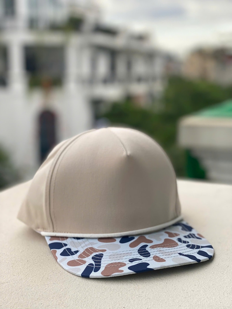 May include: A beige baseball cap with a flat peak. The peak has a pattern of blue, brown, and white shapes. The cap is made of a smooth material and has a classic, sporty design.