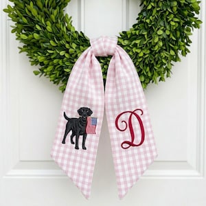 Op de afbeelding: Een roze en witte gingham lint met een zwarte Labrador Retriever met een Amerikaanse vlag en een rode geborduurde letter "D". Het lint is bevestigd aan een groene krans, waardoor een patriottisch decoratief accent ontstaat.