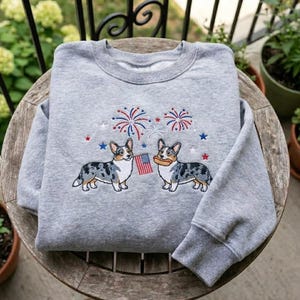 May include: A gray sweatshirt featuring an embroidered design of two corgi dogs, fireworks, and stars in red, white, and blue. One dog holds an American flag, and the other holds a hot dog. The sweatshirt has a ribbed collar, cuffs, and hem.