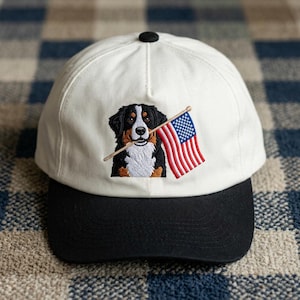 May include: White baseball cap with a black brim and a black button on top. The cap features an embroidered image of a Bernese Mountain Dog holding a small American flag in its mouth.