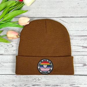 May include: A brown knit beanie with a folded brim. The beanie features a circular patch with the text "YOU'RE SAFE TO BE YOU AROUND ME" around a heart symbol in rainbow colors. The hat is on a light wood surface.