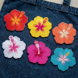 May include: Embroidered hibiscus flower patches in red, yellow, orange, white, pink, and blue. Each patch features detailed stitching and a vibrant colour palette. The patches are arranged on a denim background.