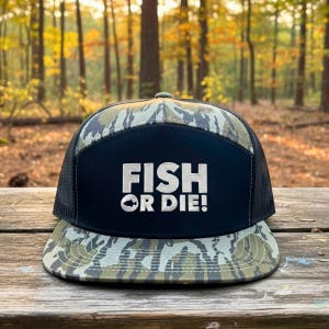Puede incluir: Gorra de camionero con estampado de camuflaje, con la parte trasera de malla negra y un panel frontal azul marino. El panel presenta el texto blanco "FISH OR DIE!" con un gr&aacute;fico de pez. La gorra est&aacute; sobre una superficie de madera.